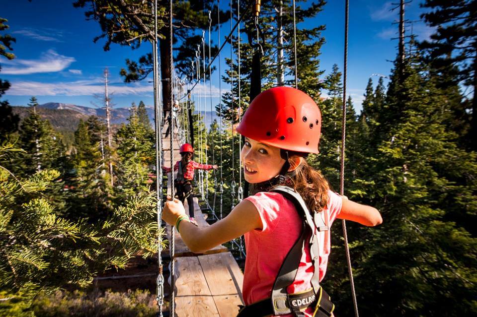 A girl wearing a harness an helmet, walking across wooden planks in the trees.